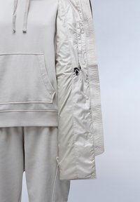 Světlounce šedá mikina s kapucí a kapsou, doplněná o sladěné tepláky. Částečně viditelná krémová prošívaná bunda s patentními knoflíky a lesklým povrchem.