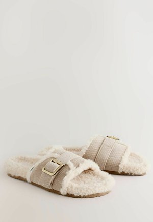 Sandales à glissière en peluche beige avec une sangle en tissu texturé et un accent de boucle dorée. Intérieur doux et moelleux et semelle plate texturée.
