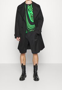Un abrigo trinchero negro sobre una camisa con patrón de remolino verde y negro. Pantalones cortos negros y botas negras de tobillo con tiradores completan el atuendo.
