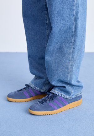 Personne portant un jean bleu et des baskets bleu marine avec des rayures violettes et des semelles beige, debout sur un sol bleu clair.