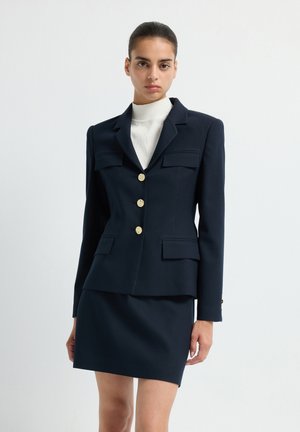 Navyblaues Blazer mit goldenen Knöpfen und zwei vorderen Taschen, kombiniert mit einem passenden, taillierten Rock. Texturiert mit vertikalen Streifen.