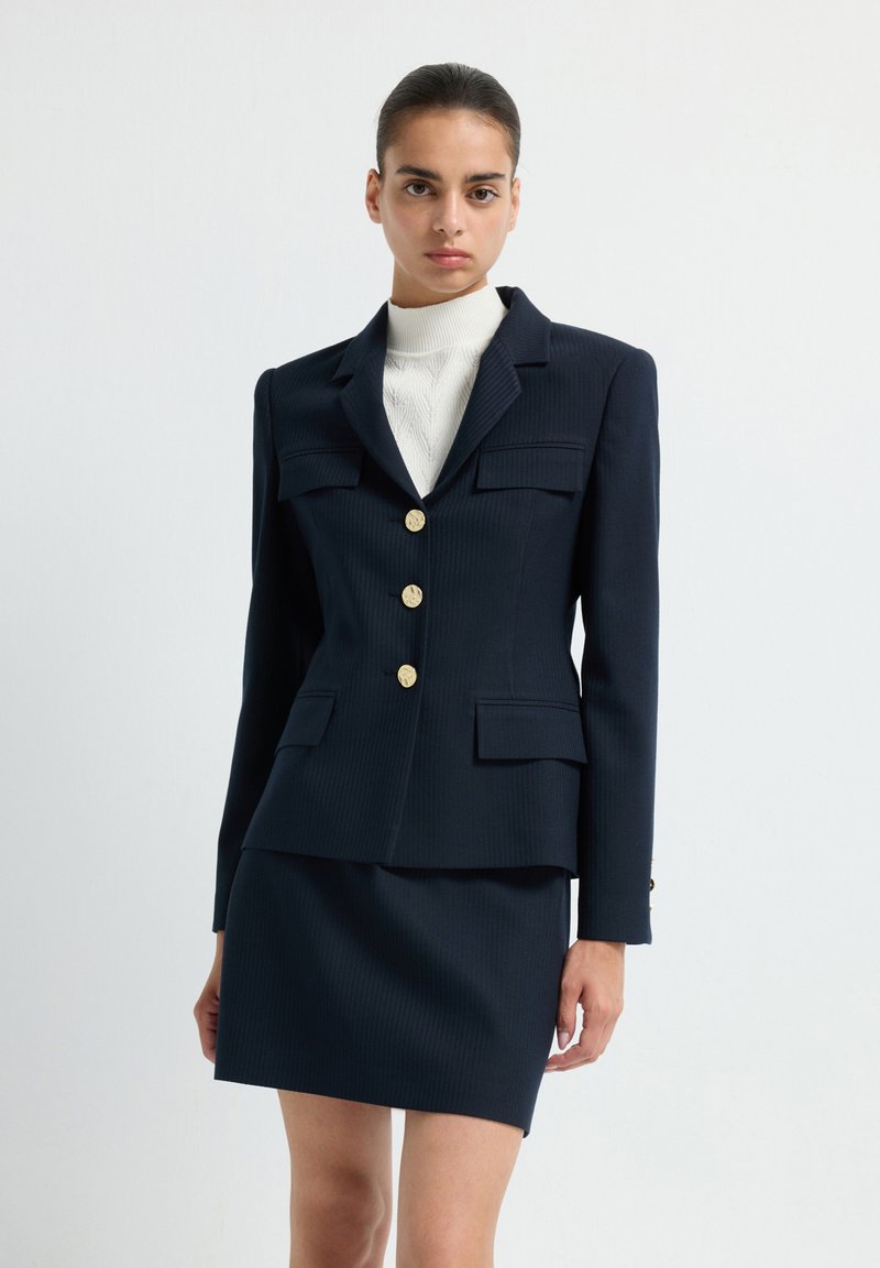 Blazer azul marino con botones dorados y dos bolsillos frontales, combinado con una falda ajustada a juego. Tejido texturizado con rayas verticales.