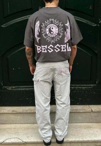 Camiseta gris de gran tamaño con un diseño gráfico rosa que presenta motivos de peces y sol, combinada con pantalones ligeros de rayas y un pequeño logo en la parte trasera.