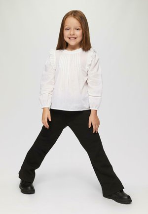 Weißes Langarmshirt mit Rüschensäumen und Falten, kombiniert mit schwarzen Schlaghosen und schwarzen Schuhen, in einer verspielten Pose stehend.