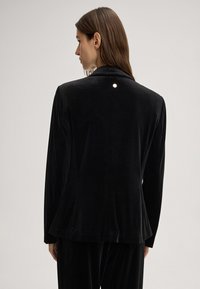 Donna con lunghi capelli castani che indossa un blazer di velluto nero e pantaloni abbinati, mostrata di spalle su uno sfondo neutro.