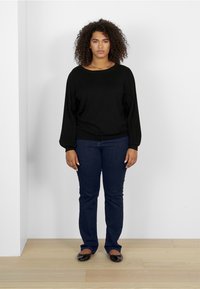 Pull en tricot noir avec des manches côtelées, associé à un jean slim bleu foncé et des ballerines noires, sur fond neutre.