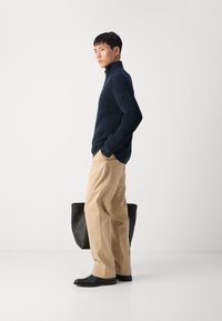 Suéter de punto acanalado en color marino con cuello alto, pantalones de pierna ancha en beige y zapatos negros. Bolsa tote negra posicionada detrás. Fondo minimalista.
