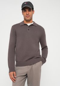 Dunkelbrauner Strickpullover mit geripptem Saum, Kragen und einer geknöpften Blende mit drei Knöpfen. Verfügt über ein kleines Logo auf der Brust und wird mit hellen Hosen getragen.