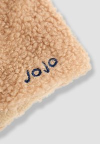 Béžová textúrovaná tkanina s nadýchaným povrchom, na okraji s tmavo modrým vyšívaným textom "Jojo". Mäkko a teplo pôsobiaci vzhľad.