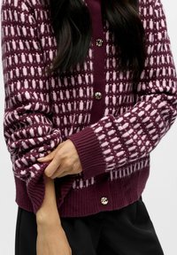 Stickad kofta i mörkröd färg med rutmönster i ljusrosa. Har ribbade muddar och en knappstängning med en metallaccent.