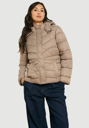 Donna che indossa una giacca imbottita trapuntata marrone chiaro con cappuccio e jeans larghi blu scuro, in piedi con le mani nelle tasche.