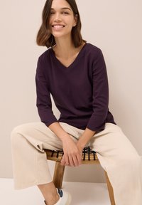 Top a manica lunga con scollatura a V in morbido cotone viola scuro. Il modello indossa pantaloni larghi beige chiaro e sneakers bianche, seduto su una sedia di legno.
