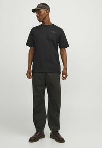 Black cotton t-shirt with short sleeves and green logo detail on left chest, paired with dark trousers and lace-up brown boots.