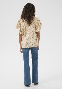 Blumige Bluse in Hellgelb mit Rüschensaum; verfügt über kleine rosa und grüne Muster. Kombiniert mit ausgestellten blauen Jeans.