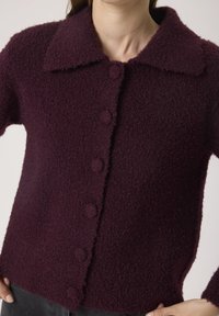 Donna che indossa un cardigan button-up di un viola scuro e texture con un ampio colletto e maniche lunghe, con le mani sui fianchi.