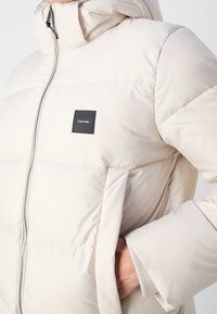 Veste matelassée beige avec fermeture éclair à l'avant, capuche, poche latérale avec main à l'intérieur, et un petit patch logo Calvin Klein noir sur la poitrine.