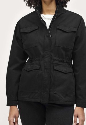 Kvinde iført en sort arbejdsjakke med fire klaplommer, høj krave og lynlås med knaplukning over en hvid skjorte.