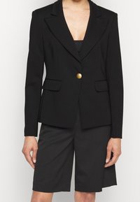 Černý blazer s jedním zlatým knoflíkem, dobře střižený design, dvě přední kapsy, hladká tkanina, klopové límce, kombinovaný s černými šortkami.