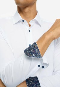 Weißes Langarmhemd mit gemustertem marineblauem Ärmelbündchen. Die Knöpfe sind marineblau, mit einer glatten Stofftextur. Verstellbare Manschetten mit Details.