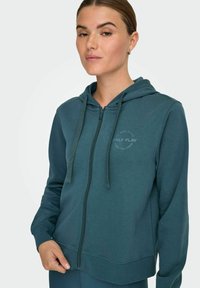 Turkizna zip-up kapuca s kapuco na vrvico. Vsebuje okroglo logo na prsih, rebraste manšete in mehak material iz bombažne mešanice.