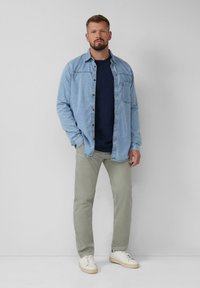 Lichtblauwe denim overhemd over een marineblauw T-shirt, gecombineerd met lichtgroene broek en witte sneakers. Minimalistisch ontwerp, casual pasvorm en zachte textuur.