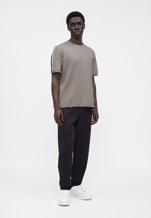 Jeune homme debout portant un t-shirt beige à manches courtes, un pantalon noir et des baskets blanches sur un fond blanc uni.