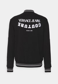 Černá mikina s bílým logem „Versace Jeans Couture“ vytištěným vzhůru nohama na zádech. Má žebrované manžety a spodní lem s bílými pruhy.