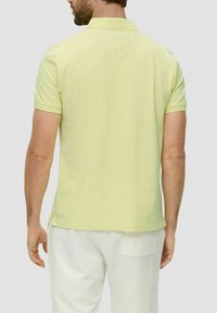 Polo-Shirt aus hellgelbem Baumwollstoff, kurze Ärmel, gerippter Kragen, gerader Saum, mit Seitenschlitzen. Getragen mit hellen Hosen.