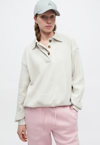 Pulover texturat de un bej deschis cu guler și trei nasturi maro, purtat cu pantaloni de trening roz pal, cu șnururi și accente cu logo.