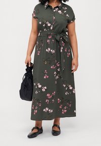 Blommig grön maxikjol med korta ärmar, knapplist framtill och midjeband. Har rosa och vita blommor, tillsammans med svarta platta skor.