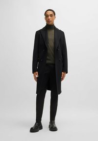 Cappotto doppio petto nero in lana, con revers a punta. Indossato sopra un dolcevita verde scuro, abbinato a pantaloni neri e stivali.