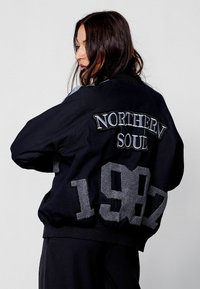 Must musta jakki hallide detailidega, mille seljal on brodeeritud "NORTHERN SOUL" ja aastaarv "1987" julgete tähtedega.