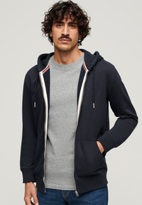 Marineblå zip-up hoodie med snoretræk i hætten, der har en grå t-shirt under. Indeholder ribbede manchetter og en kontrastfarvet indvendig foring.