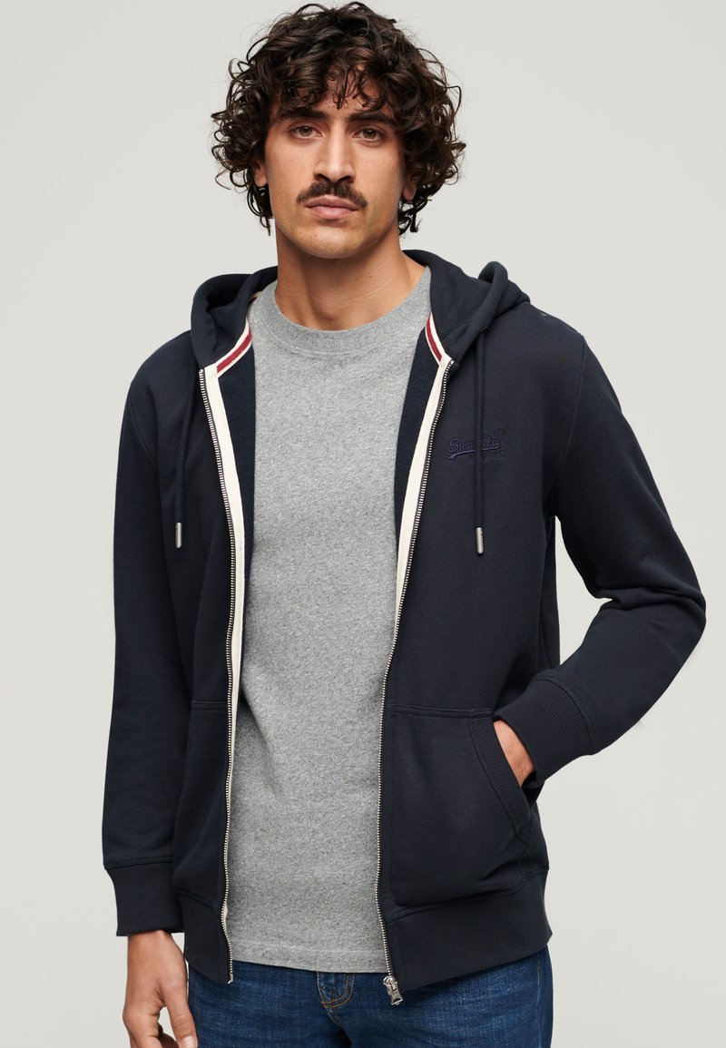Marineblå zip-up hoodie med snoretræk i hætten, der har en grå t-shirt under. Indeholder ribbede manchetter og en kontrastfarvet indvendig foring.