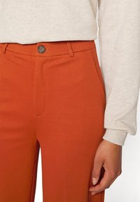 Oranžové kalhoty na míru s hladkým povrchem, které mají jednu přední kapsu a hnědý knoflík v pase. Viditelný béžový top s dlouhým rukávem.