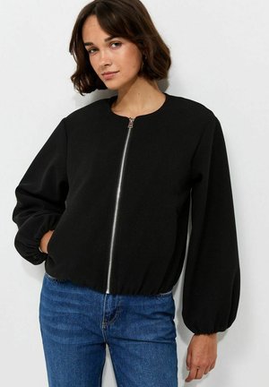 Jeune femme aux cheveux courts et bruns portant une veste noire zippée à manches bouffantes et un jean bleu sur fond blanc.