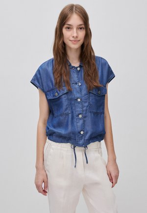 Junge Frau mit langen braunen Haaren, die ein ärmelloses blaues Jeanshemd mit Knöpfen und helle beige Hose vor schlichtem Hintergrund trägt.