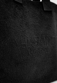 Sac fourre-tout noir en tissu doux et texturé avec une apparence duveteuse. Présente un logo en relief, des poignées robustes et un design minimaliste.