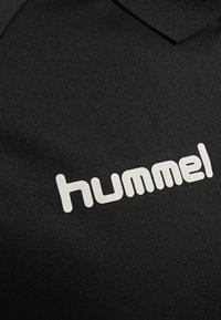 Svart pikétröja i slätt tyg med spetsig krage, med en stor vit "hummel"-logotyp på vänster sida.