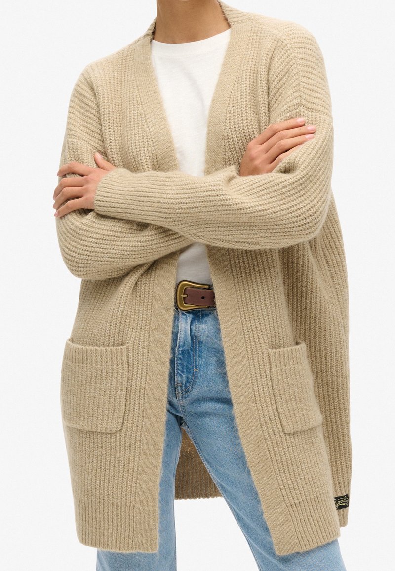 Persoon in een beige chunky gebreide cardigan met voorzakken, wit shirt, blauwe jeans en een bruine riem, met gekruiste armen.