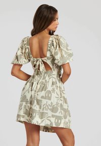 Vestido corto en beige claro con estampado floral verde, mangas abullonadas y espalda descubierta con un detalle de lazo. Diseño de falda plisada.