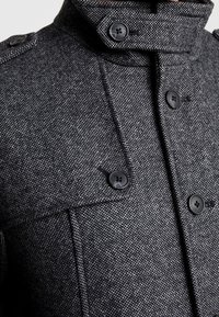 Grå texturerad kappa med hög krage, svarta knappar och detaljerad sömnad. Närbild visar tygväv och knappplaceringar.