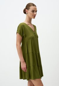 Grünes, gestuftes Kleid mit kurzen Ärmeln, V-Ausschnitt und Knopfdetails an der Vorderseite, aus einem leichten, strukturierten Stoff gefertigt.
