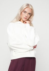 Λευκό φούτερ με καγκουρό τσέπη μπροστά, άνετη εφαρμογή και ριχτές μανσέτες. Συνδυασμένο με σκούρες μπορντό παντελόνες, σε ουδέτερο φόντο.