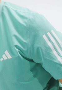 Camiseta atlética de color teal claro en tejido de malla transpirable. Cuenta con tres franjas blancas en el hombro y un logo en la parte delantera.