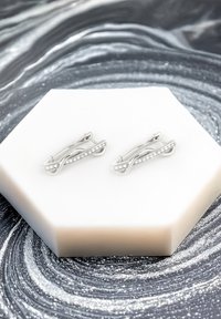 Silver clip-on örhängen i en sammanflätad design, med en rad små klara ädelstenar längs ena sidan, uppvisade på en hexagonalt vit yta.