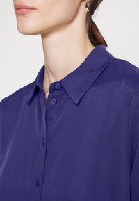 Camisa de manga curta, com colarinho, em tecido roxo profundo. Apresenta botões e um acabamento texturizado. Design simples com um corte clássico.
