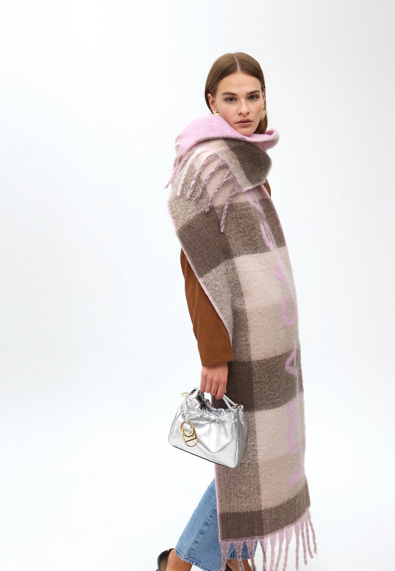 Écharpe à carreaux marron et rose avec des franges, en tissu doux. Le modèle tient un sac argenté brillant avec un cordon de serrage et des accessoires en or.
