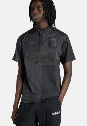 Persona vistiendo camiseta negra del Liverpool con el logo de Carlsberg y pantalones deportivos Adidas, de pie frente a un fondo blanco.