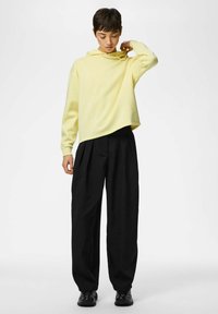 Sweat à capuche jaune raccourci avec cordon de serrage à la capuche, associé à un pantalon large noir. Le modèle se tient sur un fond uni, les mains près du col.